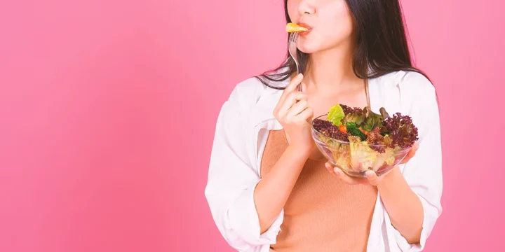 A woman eating a bowl of salad with a pink background